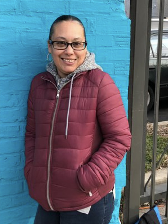 Portrait of community liaison Madelyne leaning against a blue wall and smiling