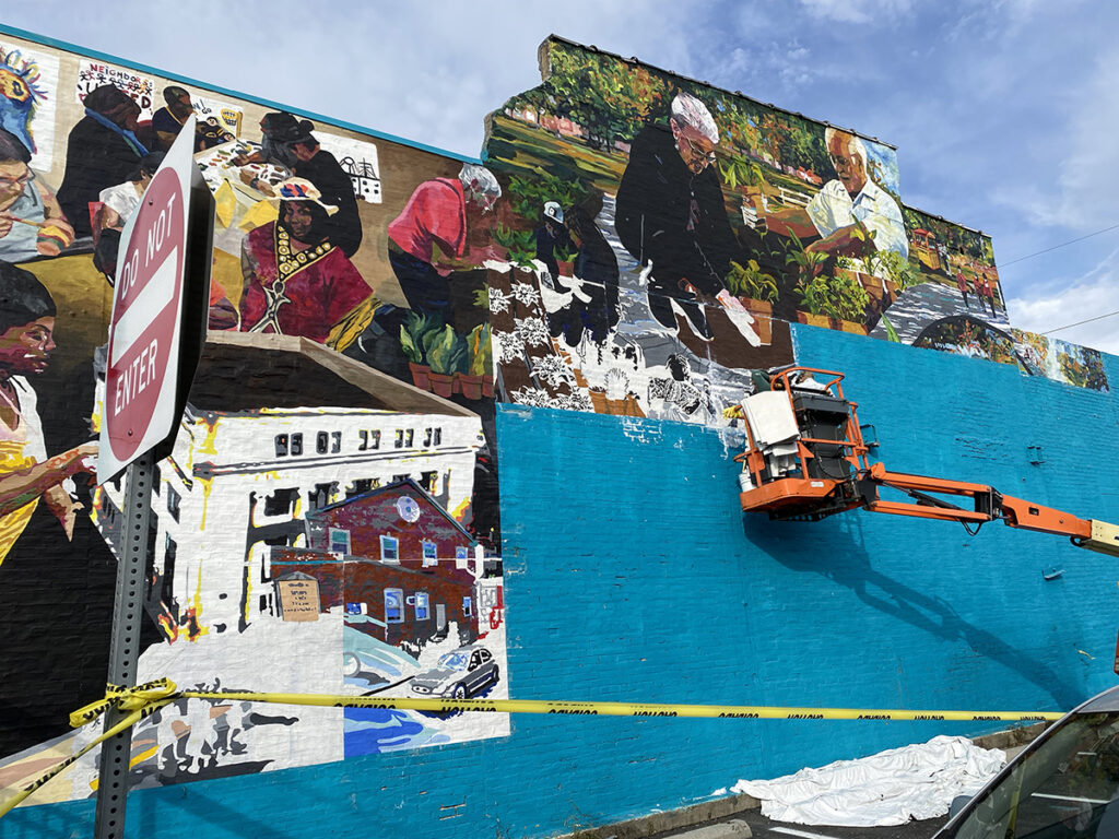 An orange forklift against a blue wall with artist Betsy Casanas adding panels to a large mural of community members