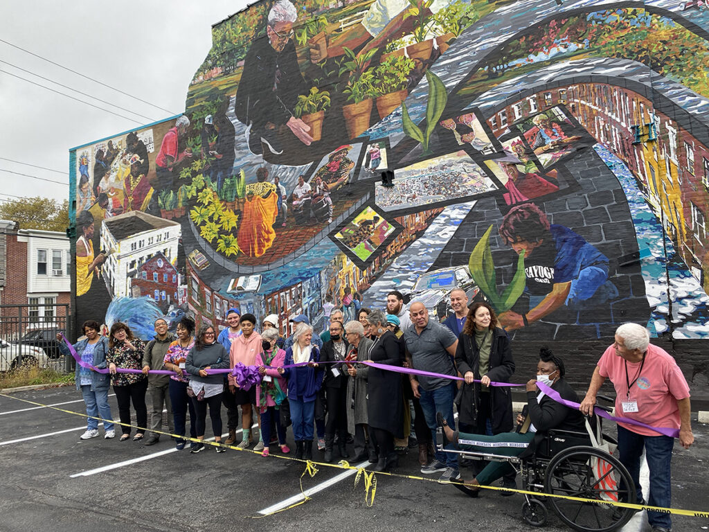A large group of community members cut a large purple ribbon in front of a colorful mural full of people and winding streets in Hunting Park
