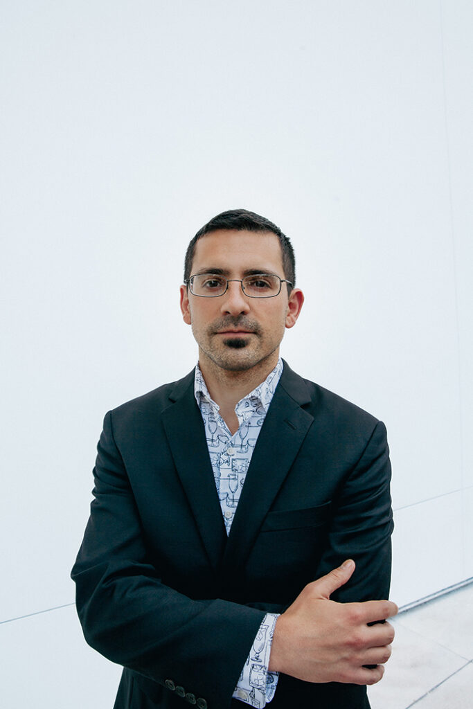 Ian Stewart waring a suit with a patterned button up standing against a white wall and looking at the viewer