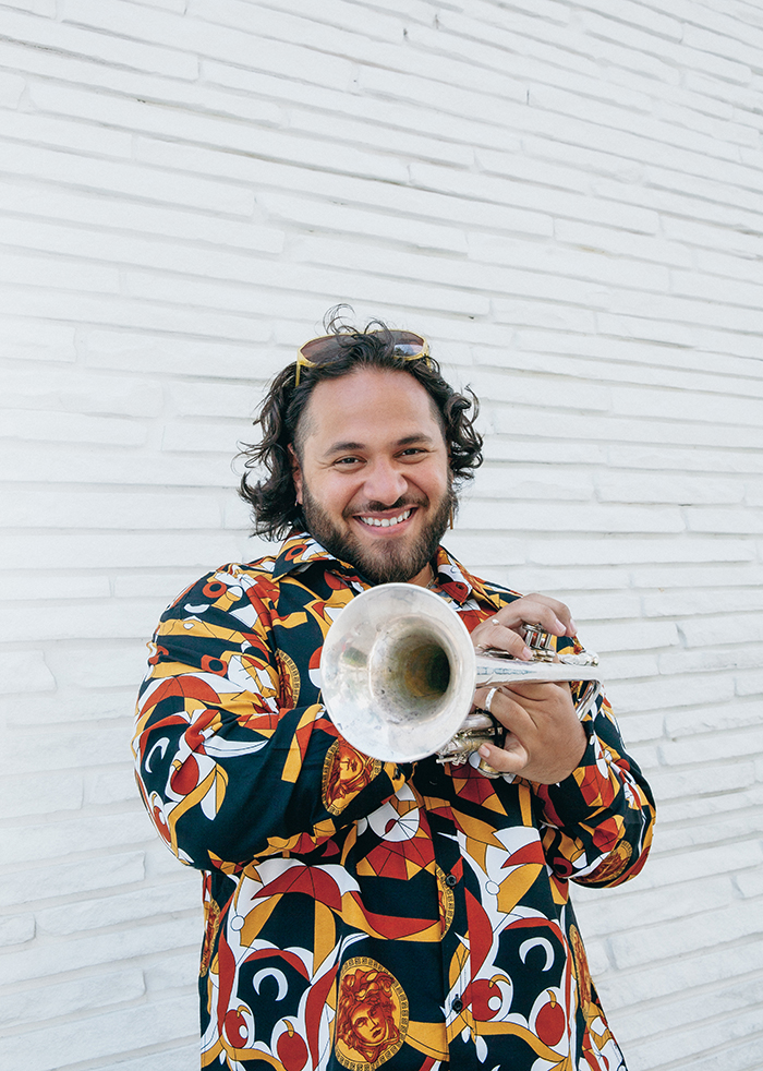 Raul Rios standing against a white wall wearing a colorful patterned button up, holding a trumpet, and smiling